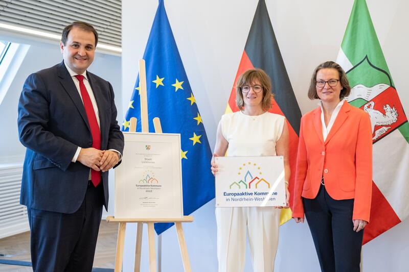 Minister Nathanael Liminski (links) und Ministerin Ina Scharrenbach (rechts) überreichen die Auszeichnung zur Europaaktiven Kommune an Bürgermeisterin Ute Dülfer (mittig)